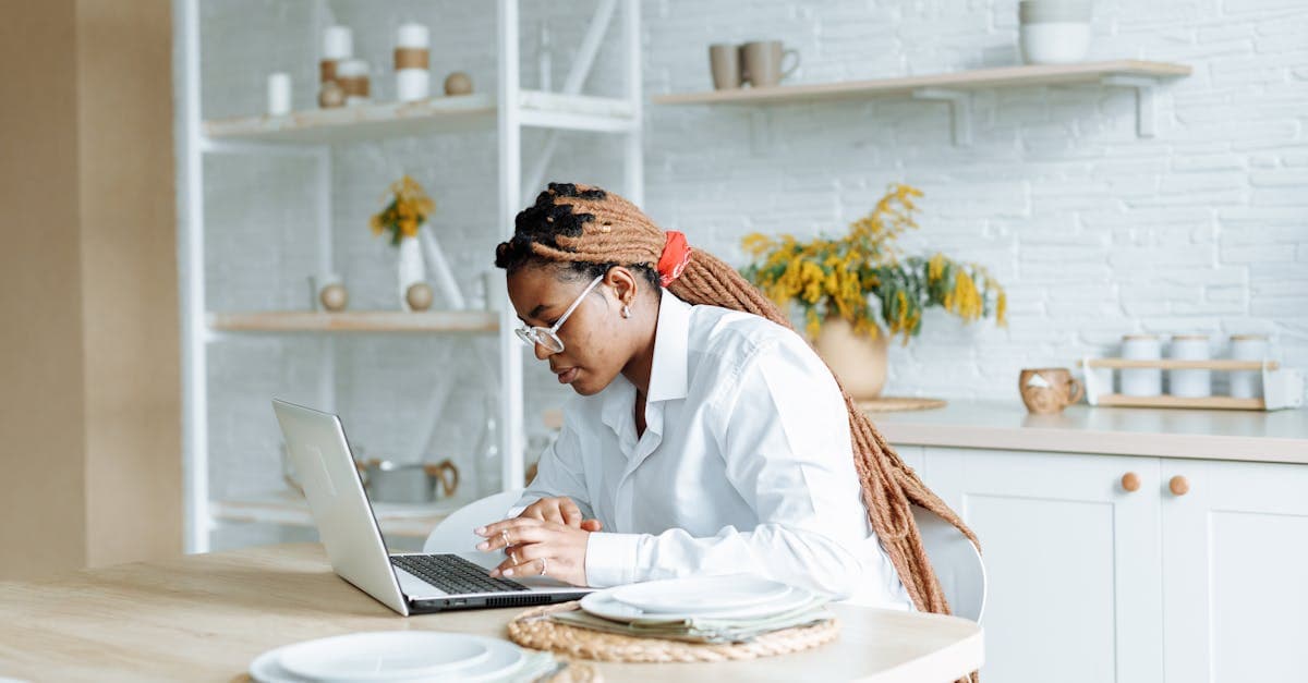 Young woman with braids working from home on a laptop in a bright, modern kitchen setting.