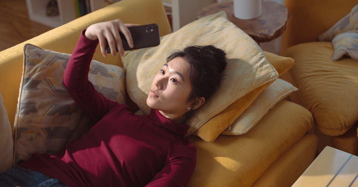Asian woman lying on a cozy sofa taking a selfie with a smartphone in a comfortable home setting.