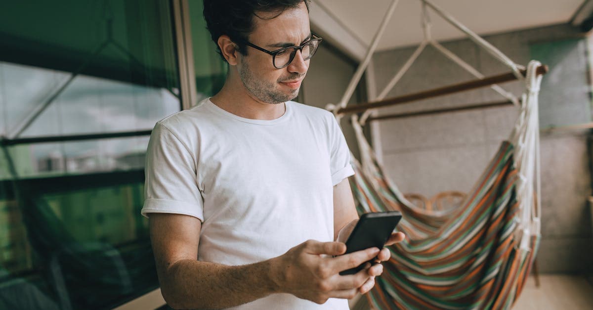 Portrait of a young man with glasses using smartphone near a colorful hammock on a balcony.