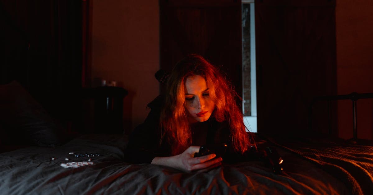 A young woman lying on a bed in a dark room, engrossed in her cellphone.