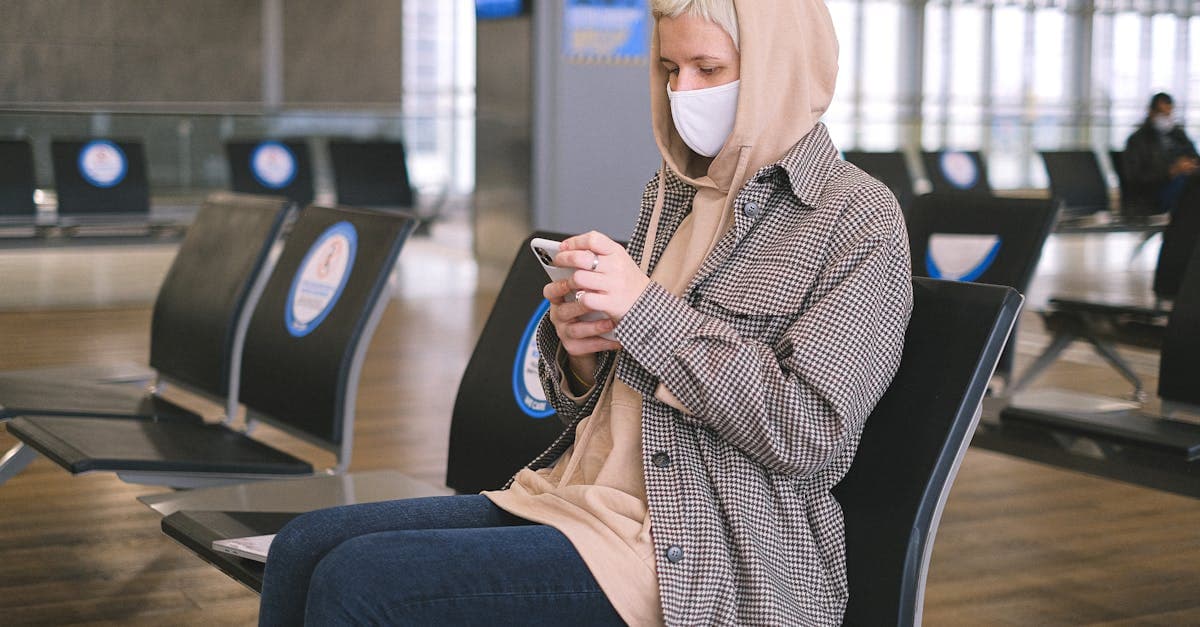 A woman wearing a face mask and a hood sits in an airport waiting area, using her smartphone.