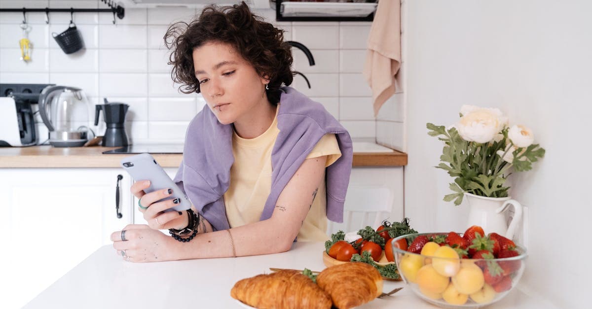 Young woman relaxing in a modern kitchen, browsing her smartphone with fresh fruits and croissants nearby.