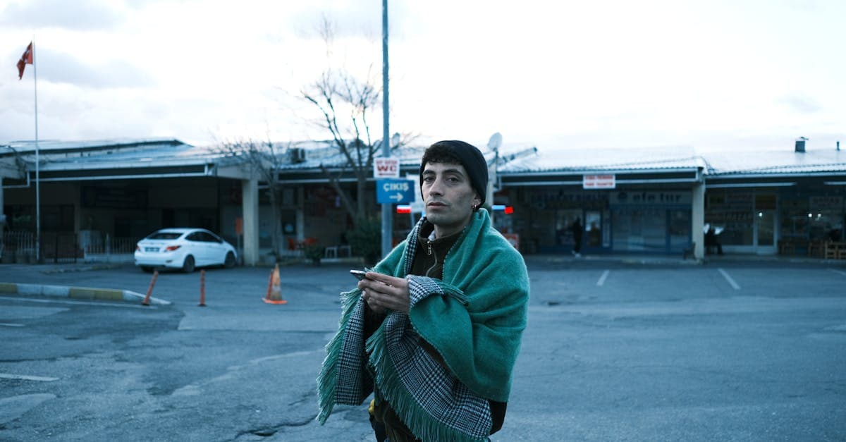 A young man stands wrapped in a blanket, holding a phone at a parking lot during dawn.