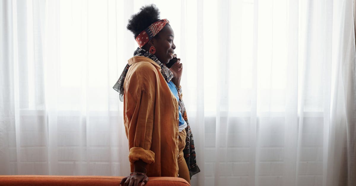 Stylish woman with headband talks on smartphone, standing by a bright window.