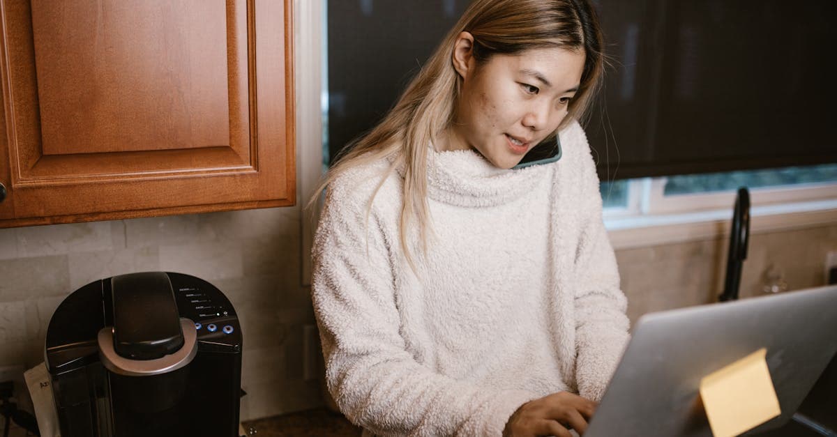 Woman multitasking on laptop and phone in a warm kitchen setup.