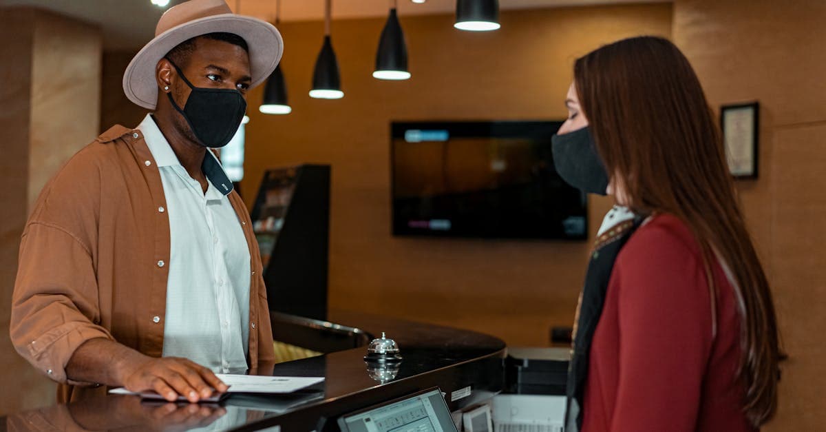 A man and woman in face masks checking in at a hotel reception, emphasizing safety protocols.