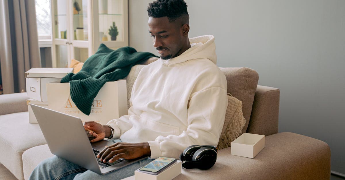 Young man in hoodie focused on laptop work at home, sitting on sofa.