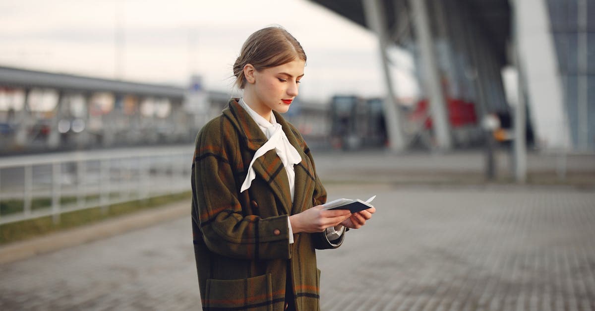 Attentive female passenger wearing trendy plaid coat and white blouse checking passport and ticket standing on pavement near modern building of airport outside