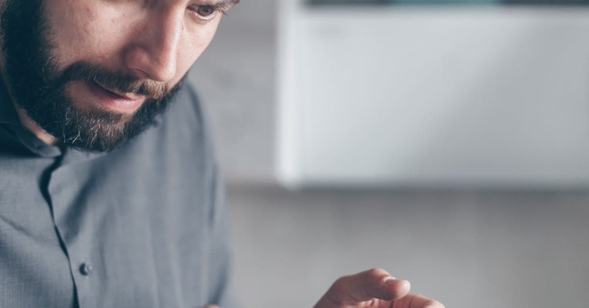 A bearded man in a gray shirt looks shocked while holding a smartphone indoors.